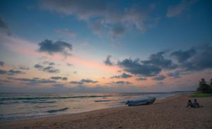 two people sitting on a beach with a boat at Cardamon Hotel Nilaveli in Nilaveli