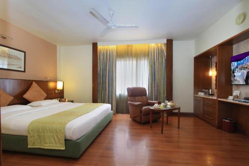 バンガロールにあるLa Sara Grandのベッド、椅子、テレビが備わるホテルルームです。