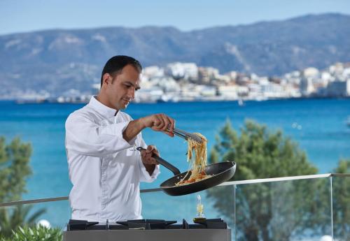 a man cooking food in a pan on a grill at Vasia Ormos Hotel (Adults Only) in Agios Nikolaos
