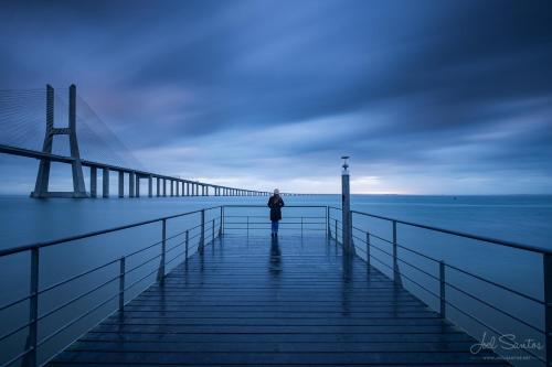 a person standing on a pier next to a bridge at A View to the River Tejo by SixtyFive Boutique Home in Lisbon