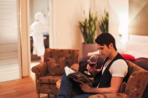 a man sitting on a couch with a book and a glass of wine at Hotel Palatina "Hotel-Restaurant" in Neustadt an der Weinstraße