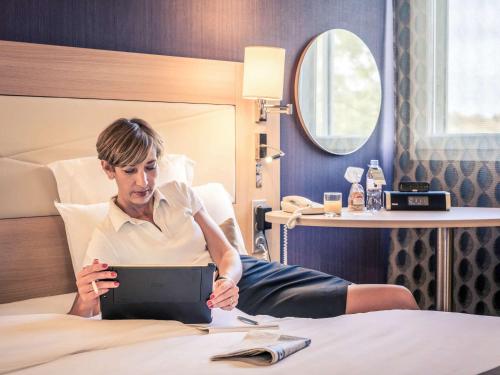 a woman sitting on a bed with a tablet at Mercure Mulhouse Centre in Mulhouse