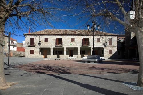 a white building with red shuttered windows and a tree at Casa Rural Soportales de Peguerinos in Peguerinos