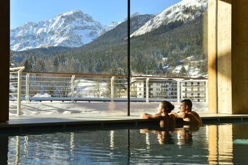 two people sitting in a swimming pool with a view of mountains at Corona Dolomites Hotel in Andalo