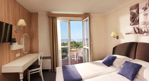 a hotel room with two beds and a desk and a balcony at Hotel Mirna - Terme & Wellness Lifeclass in Portorož