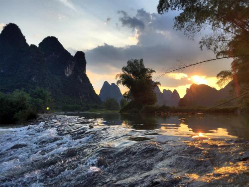 a river in front of mountains with the sun setting at The Apsara Lodge-Airport or TS Free pick-up over 3 nights in Yangshuo
