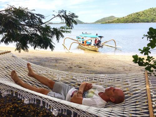 a man laying on a hammock on a beach with a boat at Palmyra Indah Bungalows in Sekotong