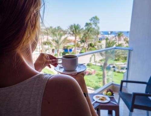 a woman holding a cup of coffee looking out the window at Seagull Beach Resort - Families and Couples Only in Hurghada