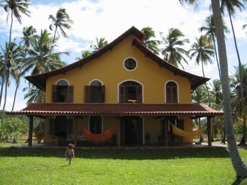 a woman standing in front of a yellow house with palm trees at Pousada Igrejinha dos Carneiros in Tamandaré
