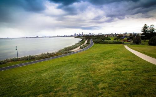 a road next to a grassy field next to the water at Beachfront Apartment in Melbourne