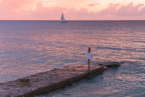a woman standing on a pier with a sailboat in the ocean at Fairmont Royal Pavilion Barbados Resort in Saint James