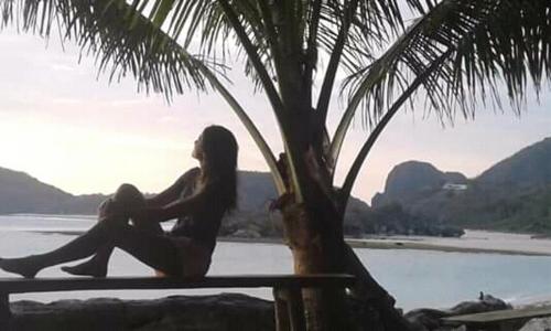 a woman sitting on a bench next to a palm tree at Castaway Surf Retreat in Sekongkang