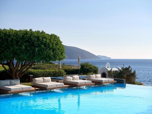 a pool with white couches and a tree and water at St. Nicolas Bay Resort Hotel & Villas in Agios Nikolaos