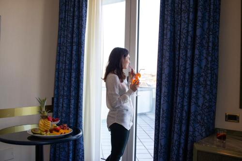 a woman standing by a window drinking a juice at Hotel Royal in Fermo
