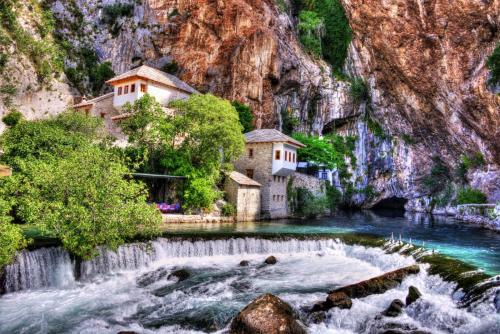 a waterfall in front of a river with buildings at Five Rivers Rooms in Mostar