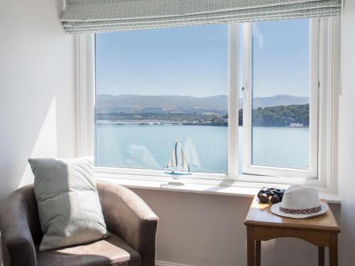 a window with a chair and a sailboat in the water at Glan Dwr Bach in Llandegfan