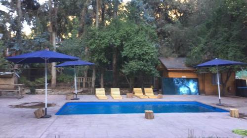 a swimming pool with two blue umbrellas and chairs at Hotel y Cabañas Rural Mirador del Maipo in San José de Maipo