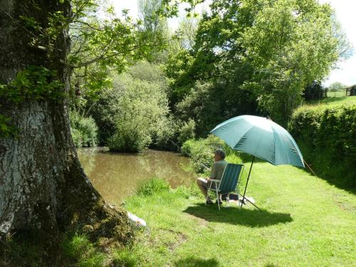 a man sitting in a chair under an umbrella next to a river at Pound Farm Holidays - Orchard Lodge in Cullompton