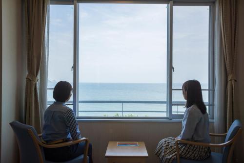 two girls sitting in chairs looking out of a window at the ocean at Kyukamura Shikanoshima in Fukuoka