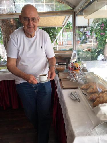 Un hombre mayor de pie junto a una mesa sosteniendo una taza. en Urcu Hotel, en Antalya