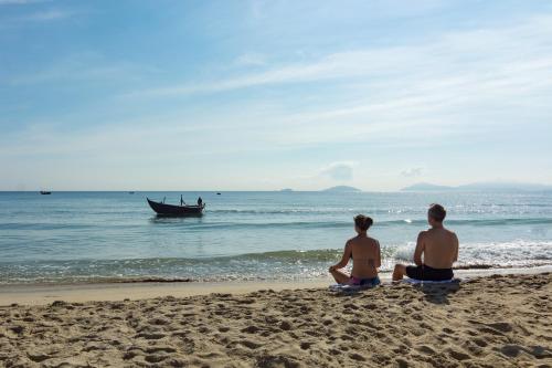 two people sitting on the beach with a boat in the water at An Bang Coco Villa in Hoi An