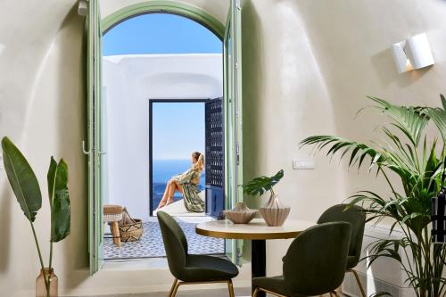 una mujer sentada en un asiento junto a la ventana en una habitación con mesa y sillas en Gitsa Cliff Luxury Villa, en Imerovigli