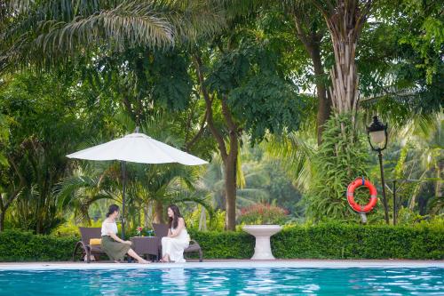 two women sitting on chairs next to a pool with an umbrella at FLC Luxury Resort Vinh Phuc in Hoằng Xá