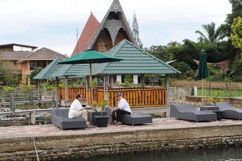 two men sitting on chairs under an umbrella near the water at Toba Village Inn in Tuk Tuk