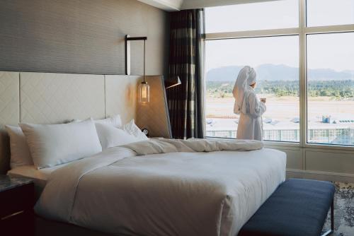 a bed in a hotel room with a woman standing next to a window at Fairmont Vancouver Airport In-Terminal Hotel in Richmond