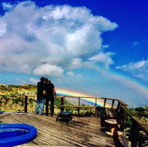 two people standing on a deck with a rainbow in the sky at El Refugio La Brisa del Diablo in Valle Hornito