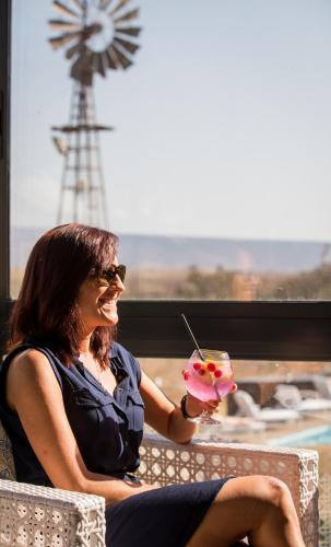 a woman holding a drink with a windmill in the background at De Zeekoe Guest Farm in Oudtshoorn