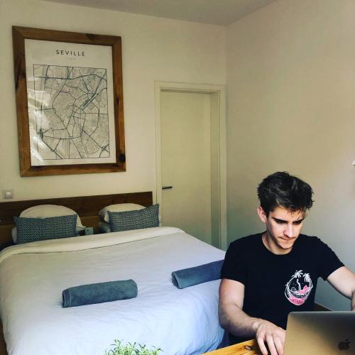 a man sitting at a desk with a laptop in front of a bed at Columbus Apartments Co-Living in Las Palmas de Gran Canaria