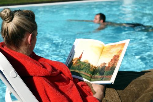 a woman sitting in a chair reading a book next to a swimming pool at Hotel & SPA Sommerfeld - Adults Only in Kremmen