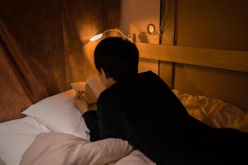a man sitting on a bed reading a book at 泊まれる図書館 寄処 -yosuga- in Toyama