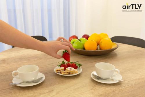a person cutting a strawberry on a table with a bowl of fruit at Localz - Sirkin Apartment in Tel Aviv