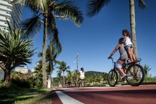two people riding bikes down a street with palm trees at Apartamento FRENTE MAR - Vista Lateral - PRAIA GRANDE in Solemar