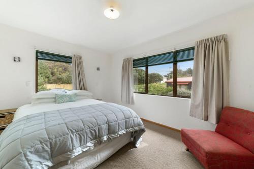 a bedroom with a bed and a red chair and windows at Heperi Haven - Waikanae Beach Holiday Home in Waikanae