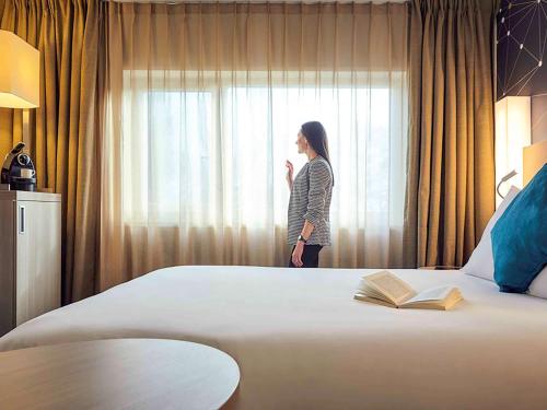 a woman standing by a bed looking out the window at Mercure Paris Sud Les Ulis-Courtaboeuf in Les Ulis
