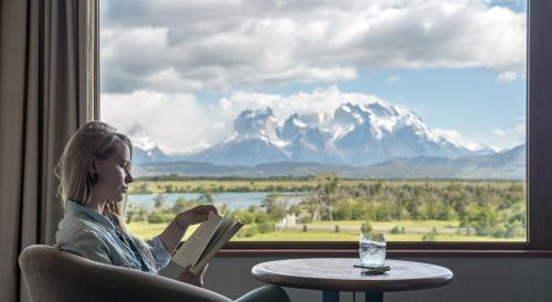 a woman sitting in a chair reading a book in front of a window at Río Serrano Hotel + Spa in Torres del Paine