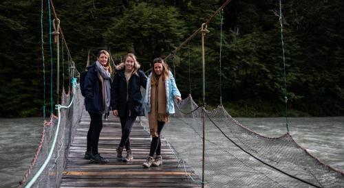 three people walking across a suspension bridge over a river at Río Serrano Hotel + Spa in Torres del Paine
