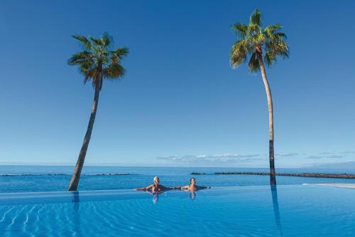 two people swimming in a swimming pool with two palm trees at Hotel Riu Arecas - Adults Only in Adeje