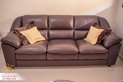 a brown leather couch with two pillows on it at MAIOR NOTA - 2 Quartos com AR CONDICIONADOS SPLIT, Air Fryer, Wi-Fi, Disney Plus, Star Plus, HBO Max, Guarda Sol, Cadeiras de Praia, Caixa Térmica, Churrasqueira, Rede para Descanso e Chuveirão - 8 pessoas - CONFORTO Máximo - Melhor Localização!! in Guarapari