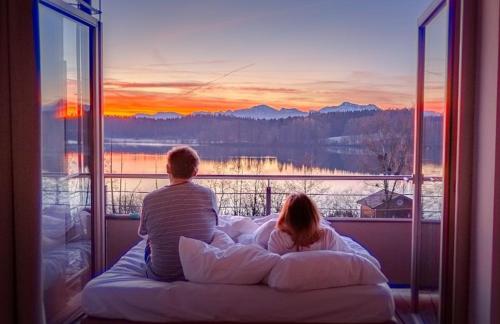a man and a woman sitting on a bed looking out a window at Lebensart am See in Bad Endorf