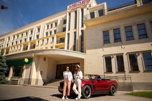 two women standing next to a red car in front of a building at Medeu in Kostanay
