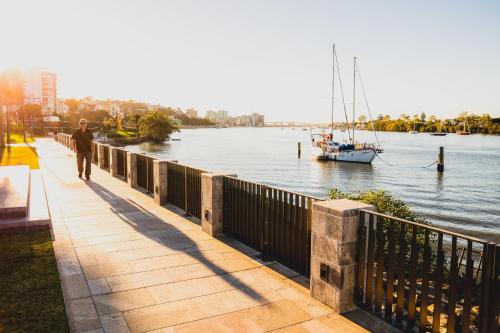 a man walking down a sidewalk next to a river with a boat at View Brisbane in Brisbane