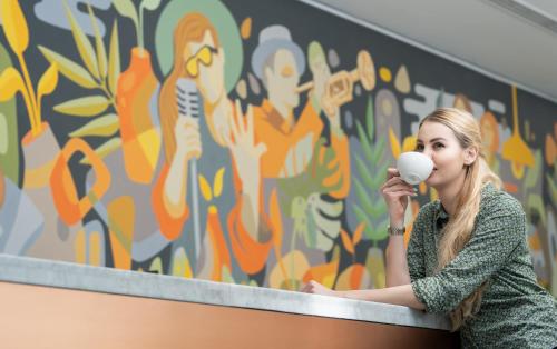 a woman drinking a cup of coffee in front of a mural at First Central Hotel Suites in Dubai