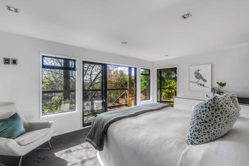 a bedroom with a bed and a chair and windows at Taonga with Sea Views by Waiheke Unlimited in Oneroa