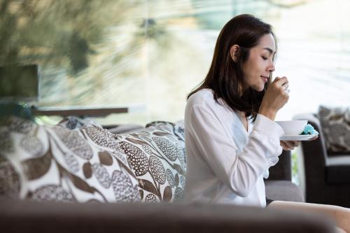 a woman sitting on a couch drinking a cup of coffee at The Centris Hotel Phatthalung in Phatthalung