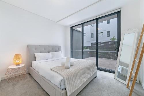 a white bedroom with a bed and a large window at KOZYGURU WAITARA KOZY 2 BED APT WALK TO TRAIN STATION NWA008 in Waitara