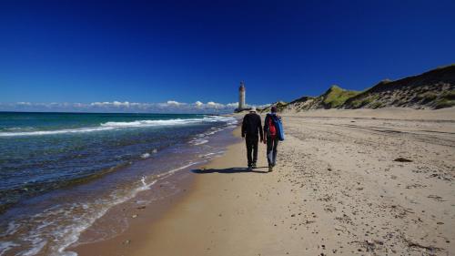 two people walking on the beach with a lighthouse in the background at Pakhuset Anholt in Anholt